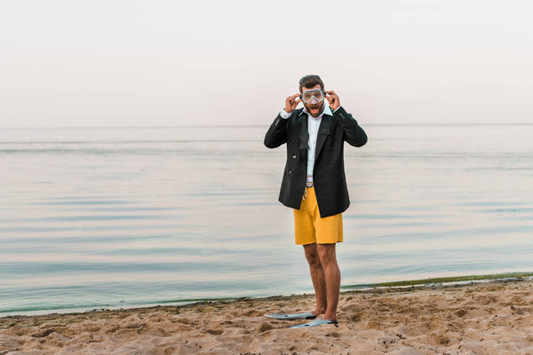 shocked man in black jacket, shorts and flippers wearing swimming mask on beach 