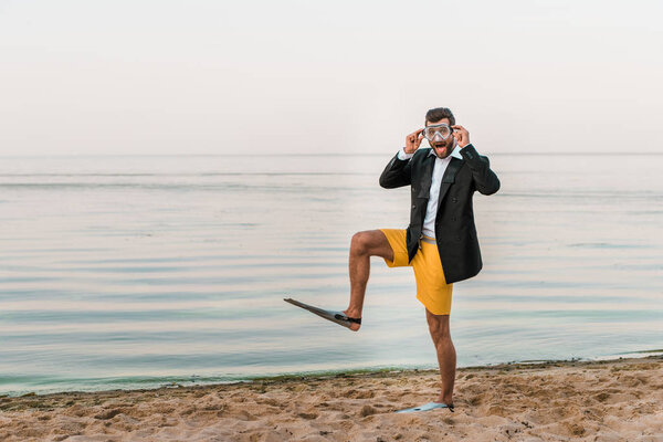 surprised man in black jacket, shorts and flippers touching swimming mask on beach near sea