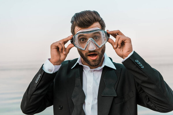 shocked man in black jacket and white shirt touching diving mask near sea