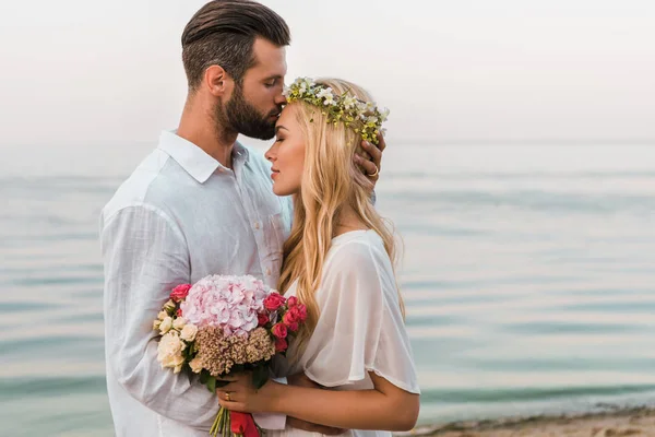 侧面观看英俊的新郎亲吻新娘额头在海滩上 — 图库照片