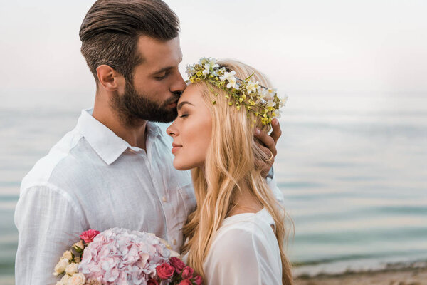 side view of handsome groom kissing attractive bride forehead on beach