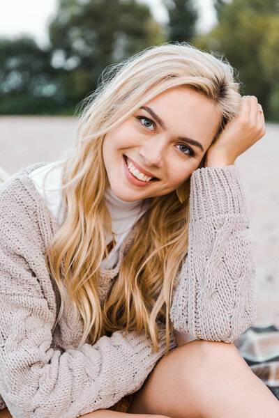 attractive smiling blonde girl in autumn outfit sitting on beach and looking at camera