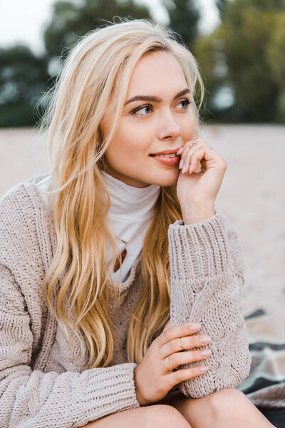 attractive smiling blonde girl in autumn outfit sitting on beach, touching lips and looking away