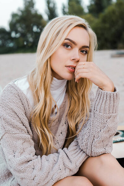 attractive blonde woman in autumn outfit sitting on beach, touching lips and looking at camera