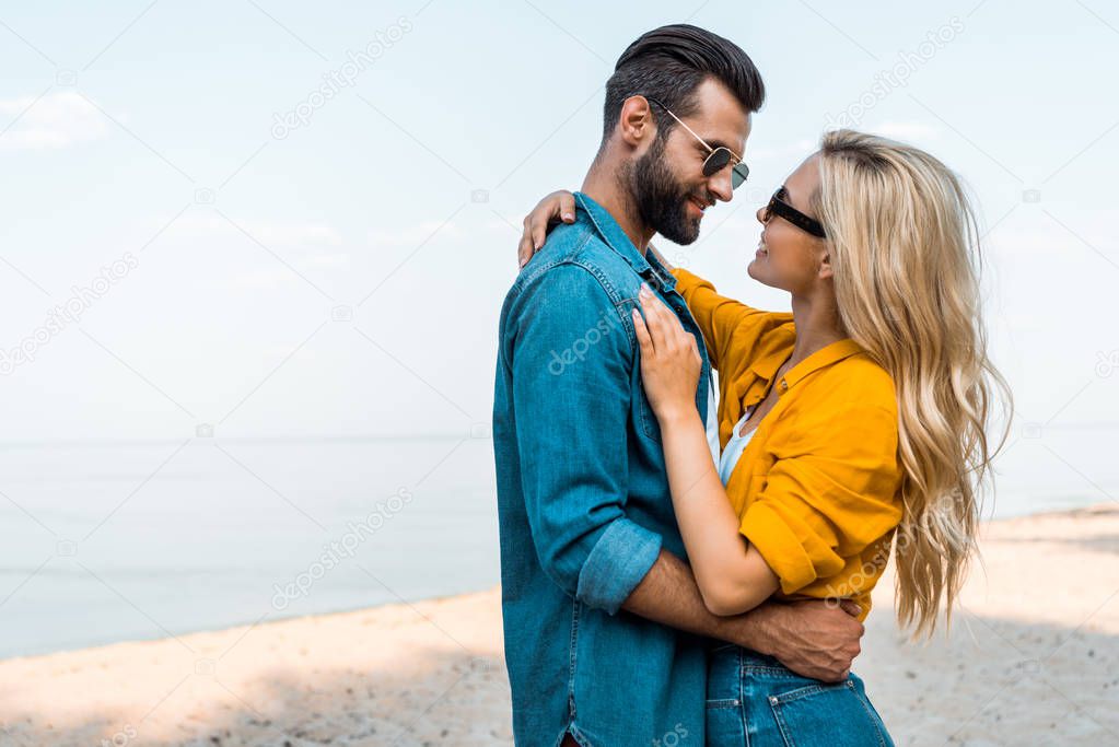 side view of couple in sunglasses hugging and looking at each other near ocean