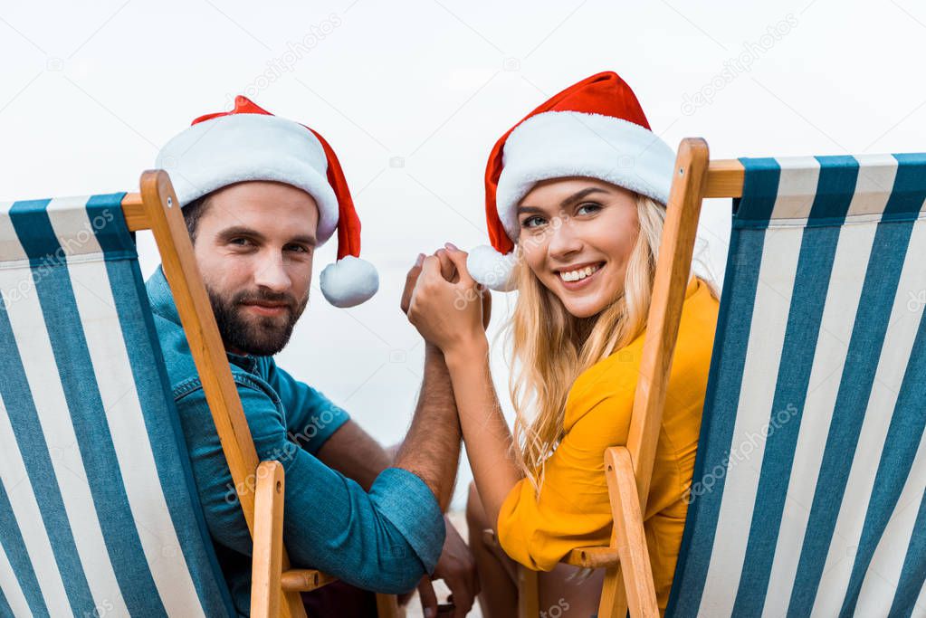 portrait of couple in santa hats sitting on sun loungers, holding hands and looking at camera on beach