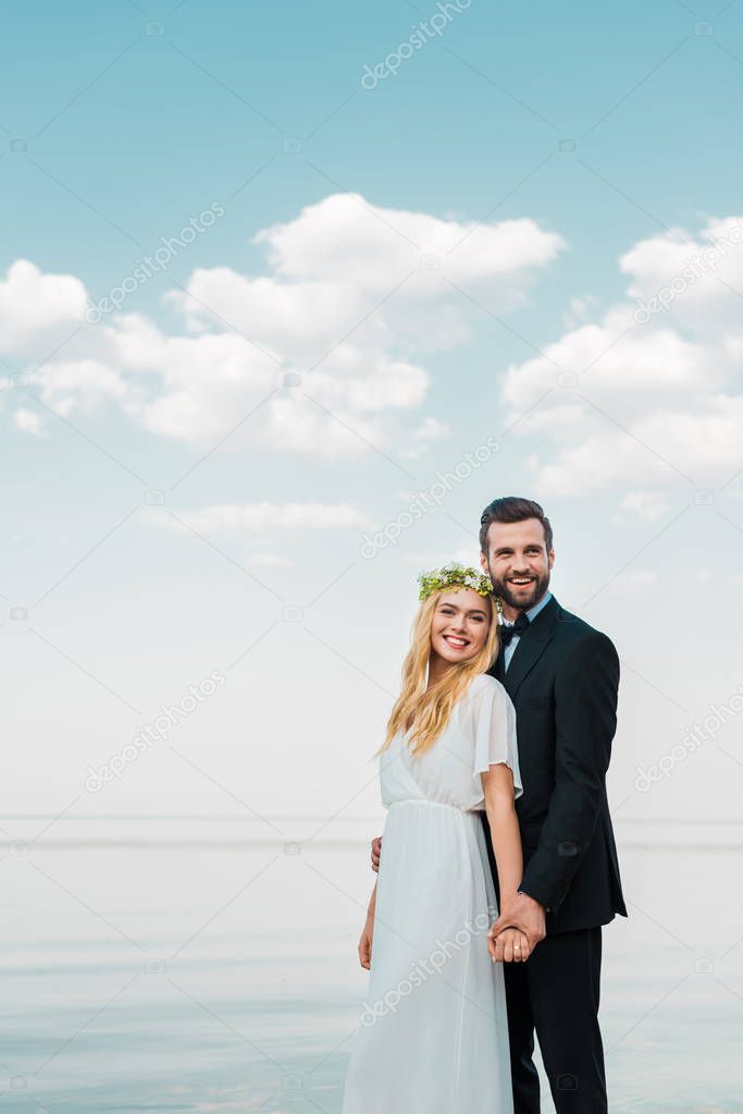 smiling wedding couple in suit and white dress holding hands on beach