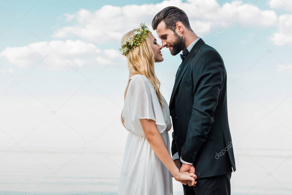 wedding couple in suit and white dress holding hands and touching with noses on beach