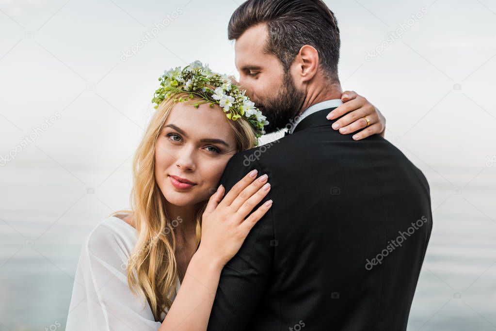 attractive bride in wreath and groom in suit cuddling on beach