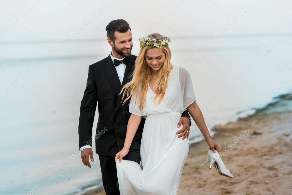 happy wedding couple hugging and walking on beach, bride holding high heels in hand