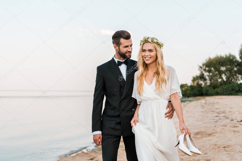 groom hugging bride and walking on beach, bride holding high heels in hand