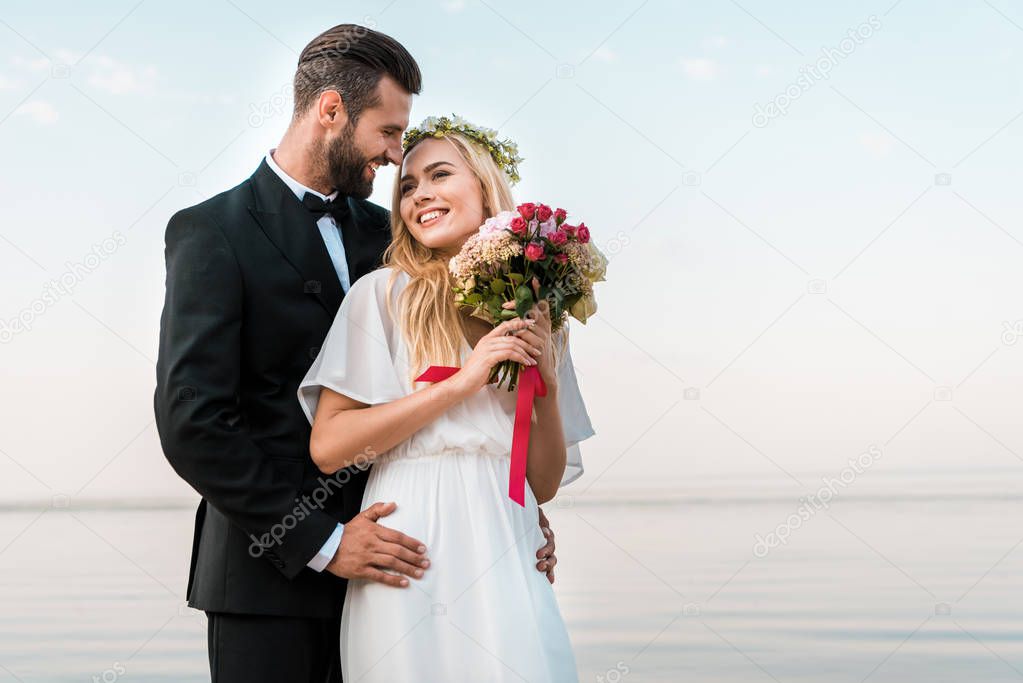 groom hugging smiling bride and she holding wedding bouquet on beach