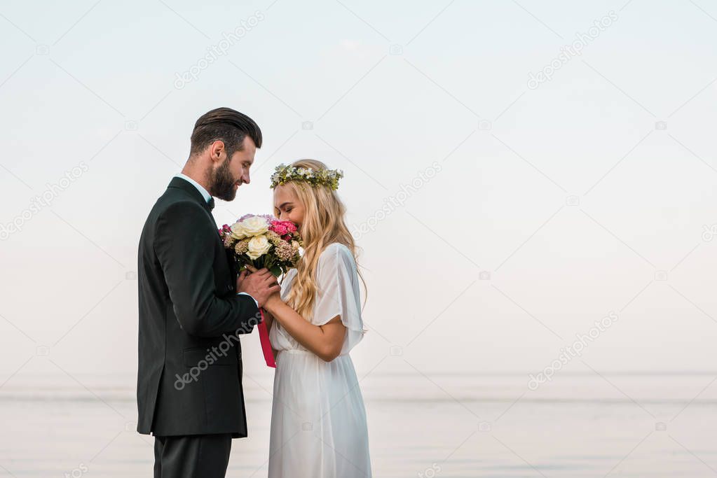 side view of wedding couple standing with bouquet on beach, bride sniffing roses