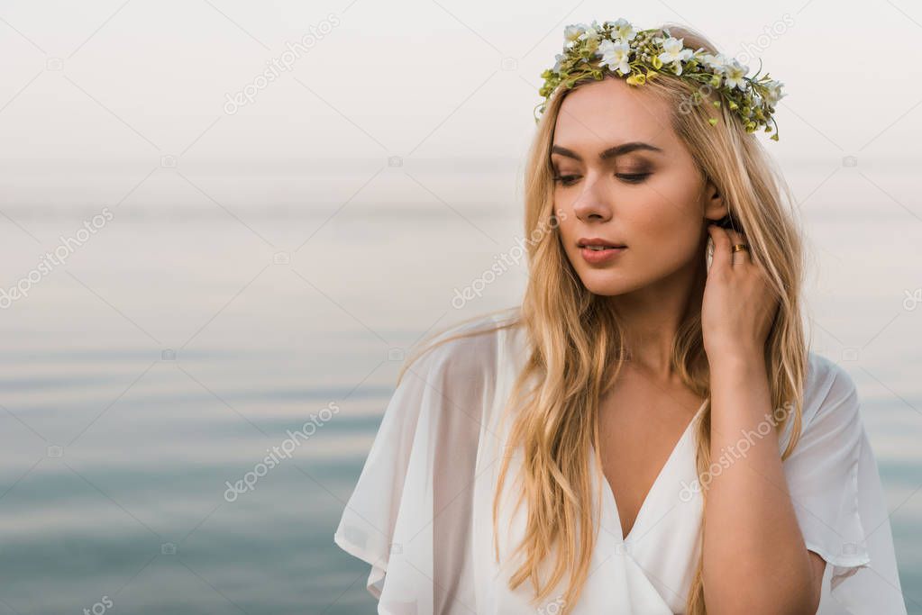 attractive bride in white dress and wreath of flowers touching hair on beach