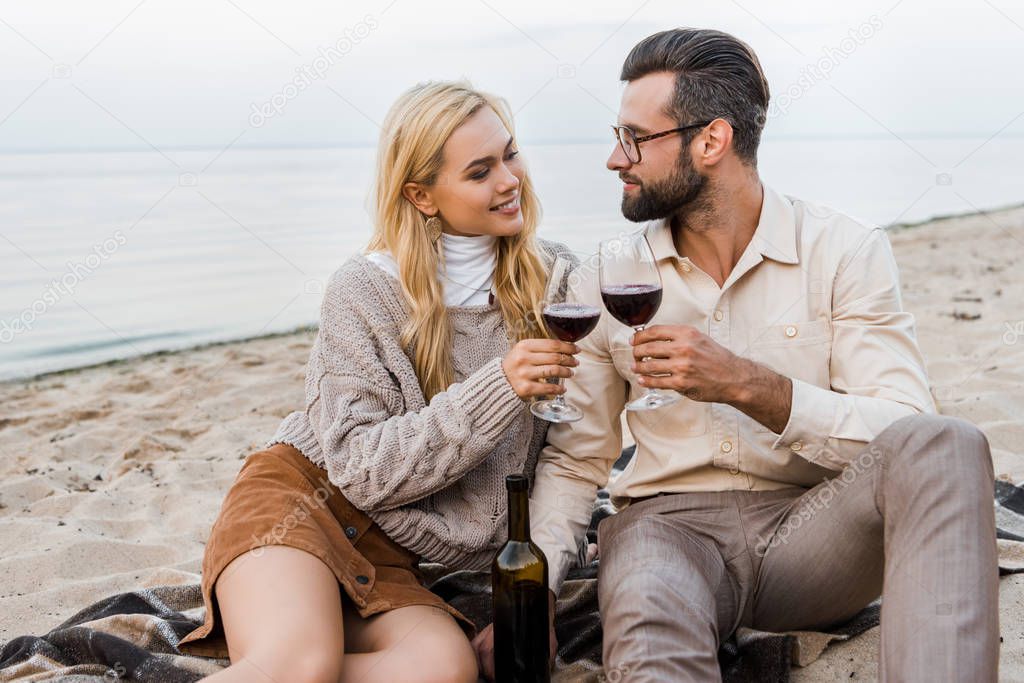 happy boyfriend and girlfriend in autumn outfit clinking with glasses on beach