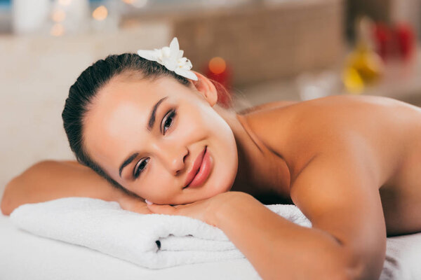 happy woman with flower in hair relaxing at spa salon and looking at camera
