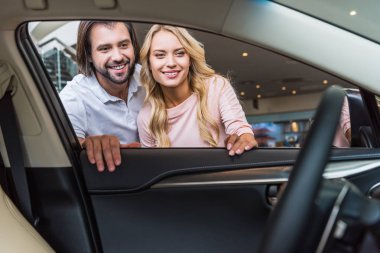 smiling couple looking into new car at dealership salon clipart