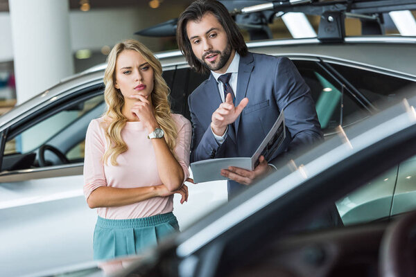 seller in formal wear recommending automobile to woman at dealership salon