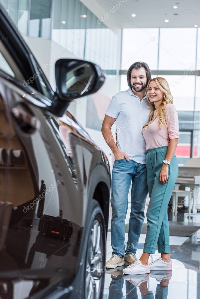 happy couple looking at new automobile at dealership salon