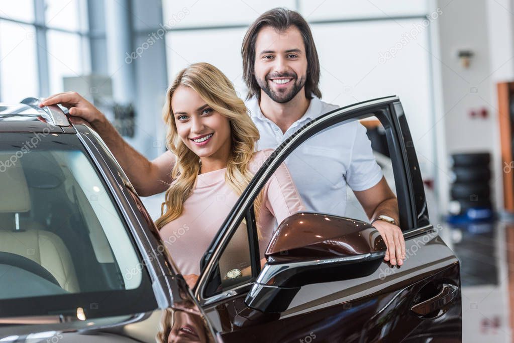 portrait of smiling couple standing at new car at dealership salon