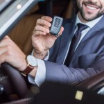 Partial view of smiling businessman with car key in hand sitting in new car in dealership salon