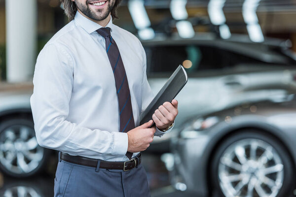 cropped shot of seller with folder standing in dealership salon with cars on background