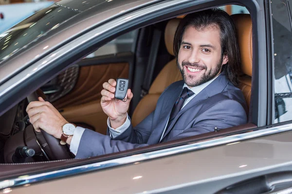 happy businessman showing car key while sitting in new car in dealership salon