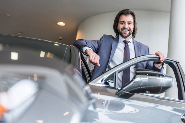 счастливый стильный бизнесмен, стоящий у нового автомобиля в салоне автосалона

