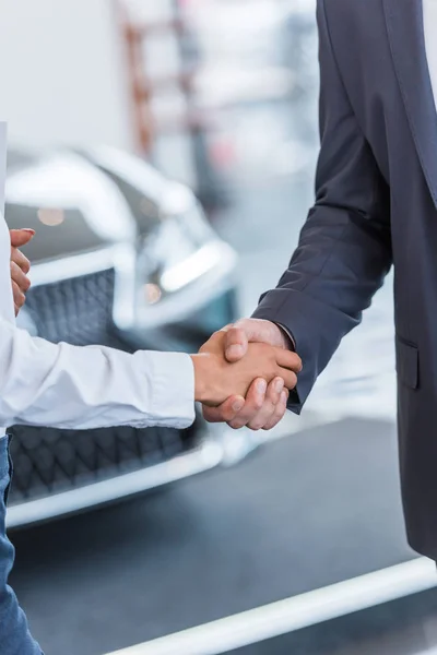 cropped shot of auto salon seller and businessman shaking hands at dealership salon
