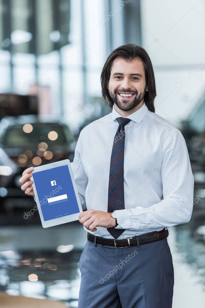 Smiling dealership salon seller in formal wear showing tablet with facebook logo on screen in hands