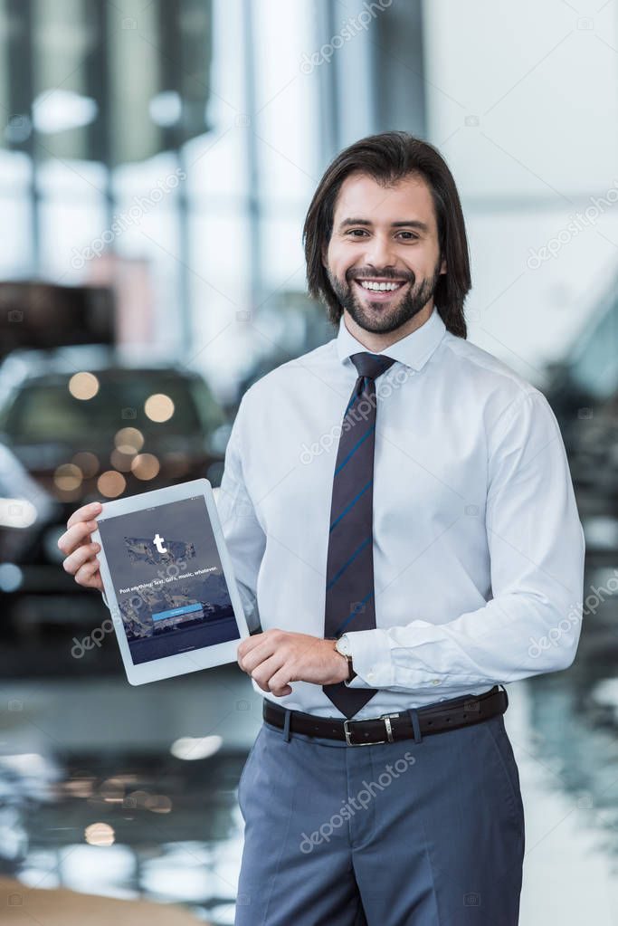 Smiling dealership salon seller in formal wear showing tablet with tumblr logo on screen in hands