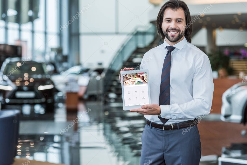 Portrait of cheerful dealership salon seller in formal wear showing tablet with foursquare logo on screen in hands