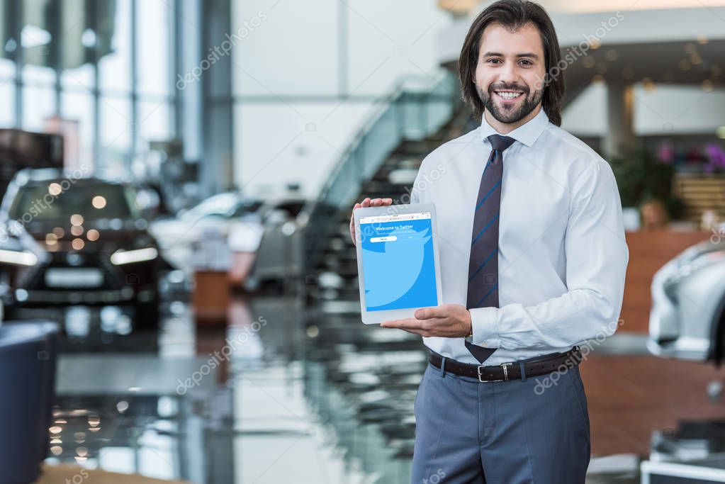 Portrait of cheerful dealership salon seller in formal wear showing tablet with twitter logo on screen in hands