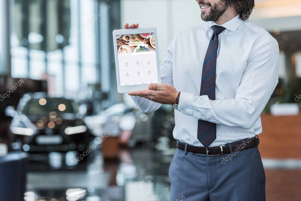 Partial view of dealership salon seller in formal wear showing tablet with foursquare website on screen in hands