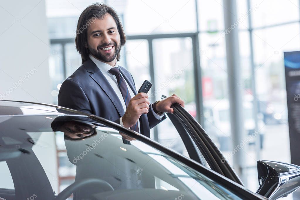 portrait of smiling businessman with car key standing at new car at dealership salon