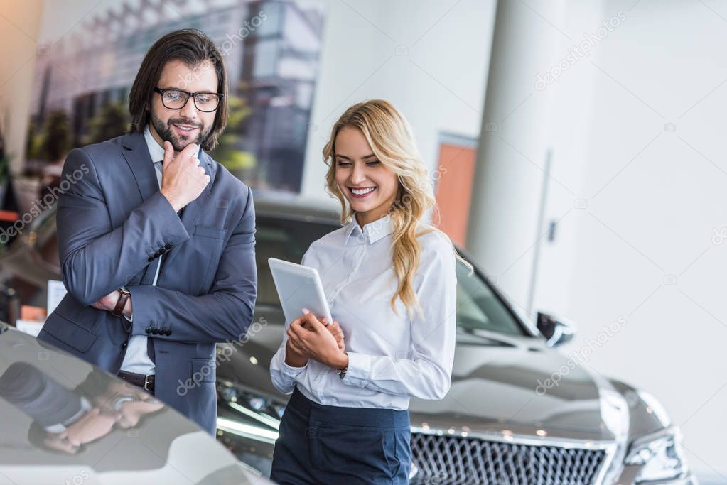 smiling female auto salon seller with tablet helping businessman to choose car at dealership salon