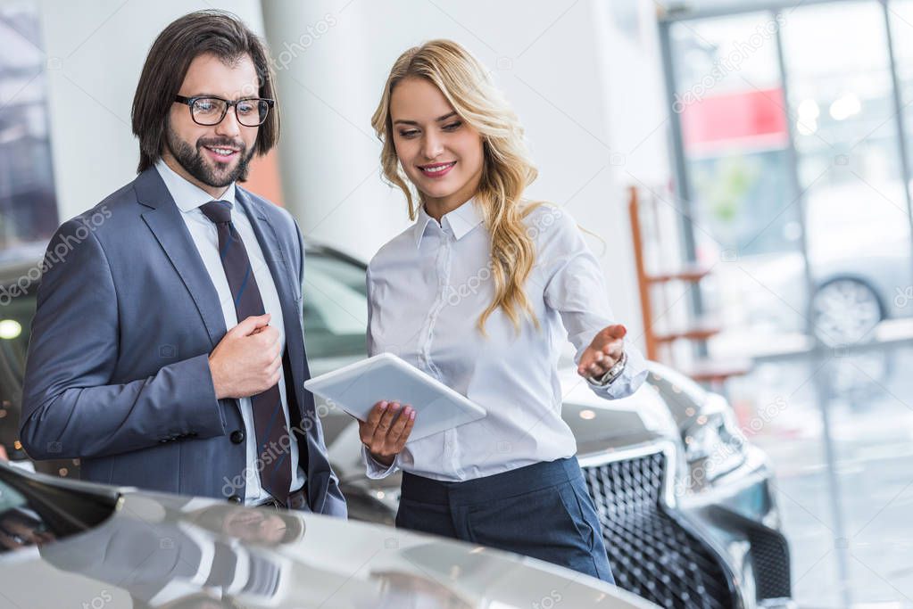 female auto salon seller with tablet helping businessman to choose car at dealership salon