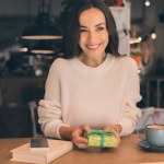 Glad ung kvinna håller presentförpackning vid bord med kaffekopp och smartphone i café