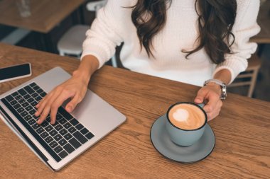 kadın Freelancer ile smartphone ve kahve Kupası kafede masada otururken dizüstü bilgisayarda çalışan kısmi görünümü 