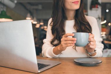 partial view of female freelancer sitting with laptop and coffee cup at table in cafe clipart