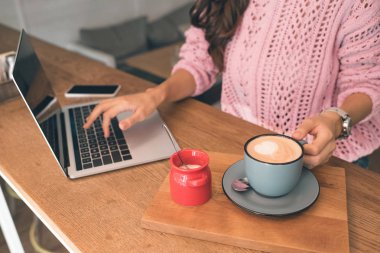 kadın Freelancer ile smartphone ve kahve Kupası kafede masada otururken dizüstü bilgisayarda çalışan kısmi görünümü 