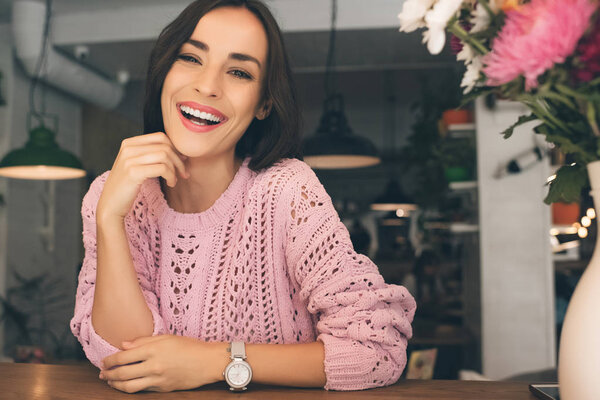 laughing pretty young woman looking at camera while sitting at table in cafe