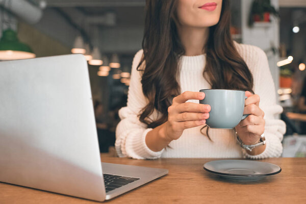 partial view of female freelancer sitting with laptop and coffee cup at table in cafe