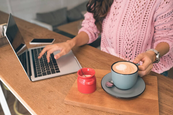 частичный вид женщины-фрилансера, работающей на ноутбуке, сидя за столом со смартфоном и кофейной чашкой в кафе
 