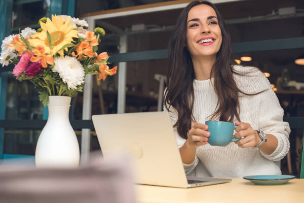 happy female freelancer with coffee cup sitting at table with laptop in cafe