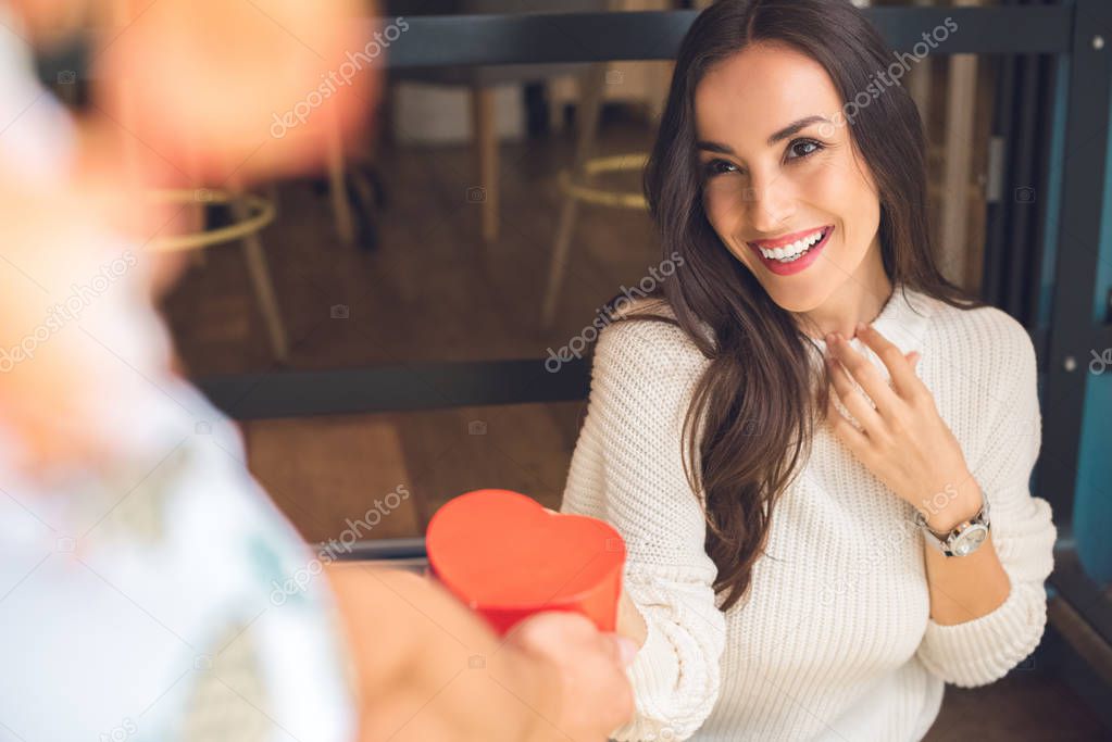 partial view of man presenting heart shaped gift box to happy girlfriend at table in cafe 