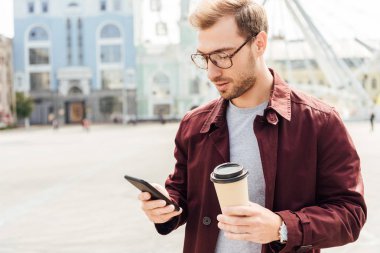 handsome man in autumn outfit holding coffee to go and using smartphone in city clipart