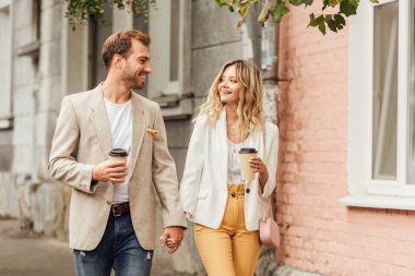 smiling couple in autumn outfit holding hands, looking at each other and walking on street with disposable coffee cups