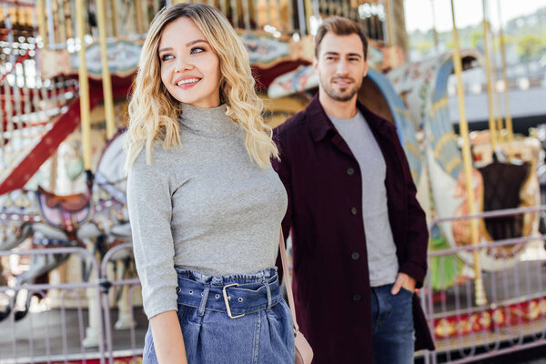 smiling affectionate couple in autumn outfit looking away near carousel in amusement park