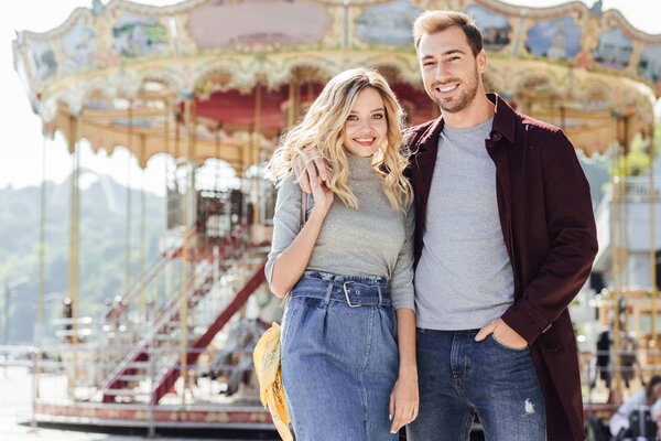 smiling affectionate couple in autumn outfit cuddling near carousel in amusement park and looking at camera